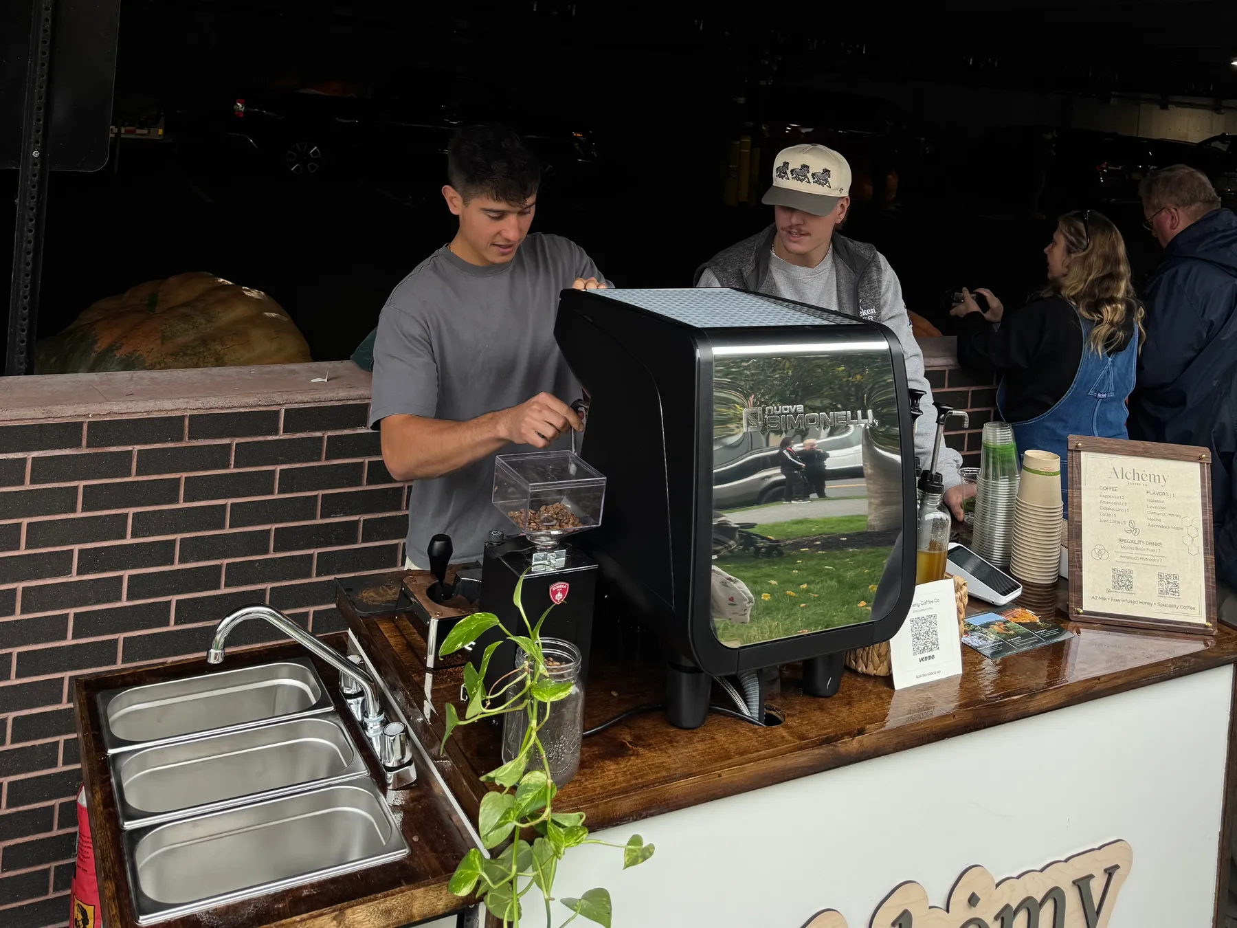 Two baristas at the Alchemy cart at an outdoor pop-up, one grinding beans beside the espresso machine, with guests waiting at the end of the counter