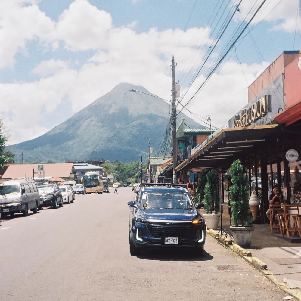 A Costa Rican street of shops and parked cars, a cone-shaped volcano rising behind
